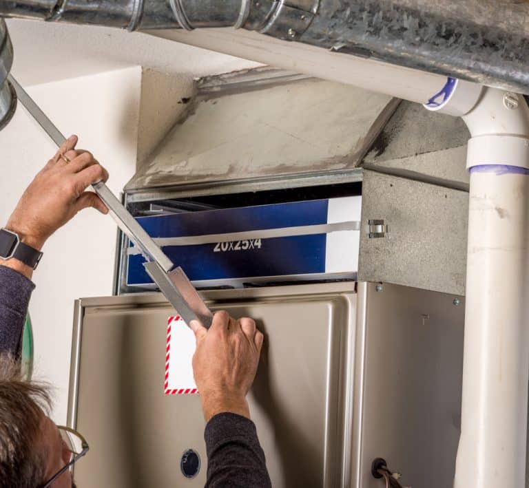 A person installs or changes an air filter in a home HVAC furnace system, sealing the compartment with duct tape. The system is mounted indoors with visible ductwork and pipes, similar to setups often seen in furnace repair by plumbers.