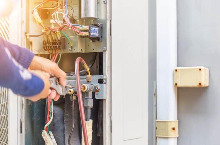 A person’s hands use a wrench to adjust pipes on an open air conditioning unit, exposing colorful wires and internal components. Sunlight shines on the scene, highlighting expert hvac work by Alpha Plumbing Heating and Cooling.