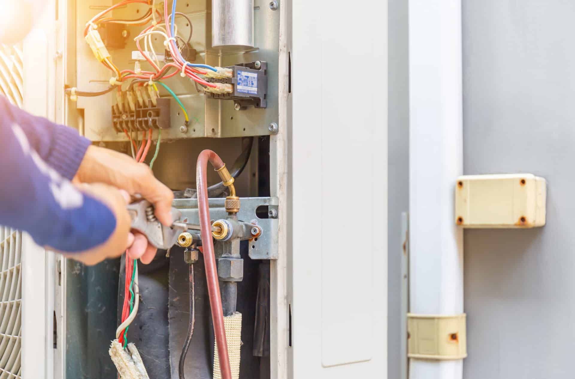 A person’s hands use a wrench to adjust pipes on an open air conditioning unit, exposing colorful wires and internal components. Sunlight shines on the scene, highlighting expert hvac work by Alpha Plumbing Heating and Cooling.