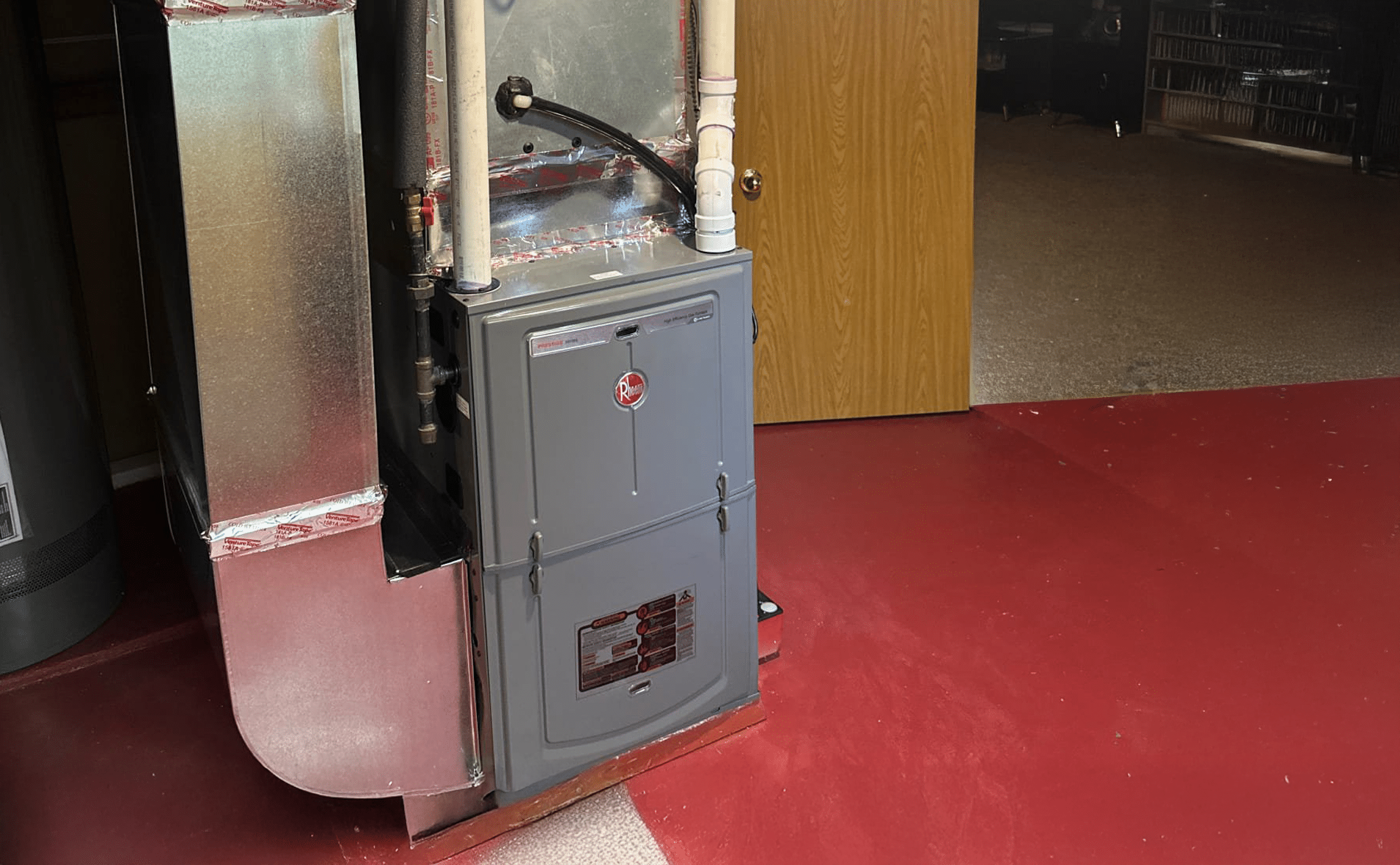 A gray Rheem furnace unit with metal ductwork, serviced by Alpha plumbing heating and cooling, is installed on a red painted basement floor near an open wooden door, leading to a carpeted room in the background.