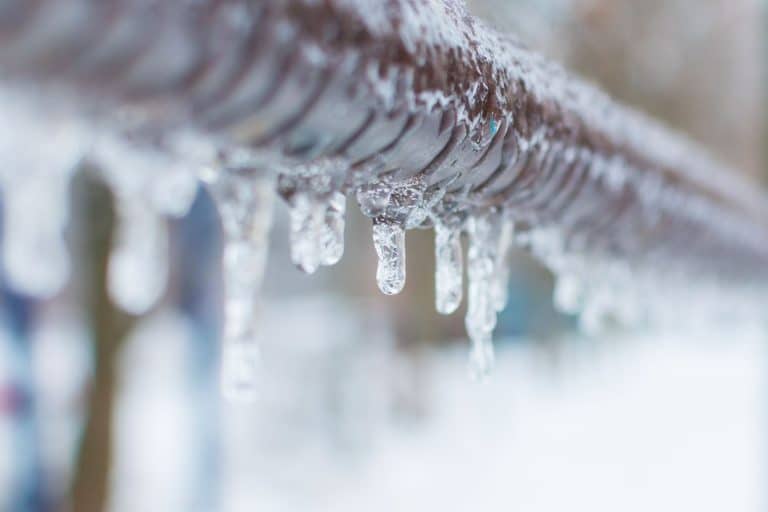 Close-up of a metal fence covered in icicles, with a snowy, blurred background. The scene captures the harsh winter weather—reminding homeowners to call Alpha Plumbing Heating and Cooling plumbers for reliable HVAC solutions during the cold.