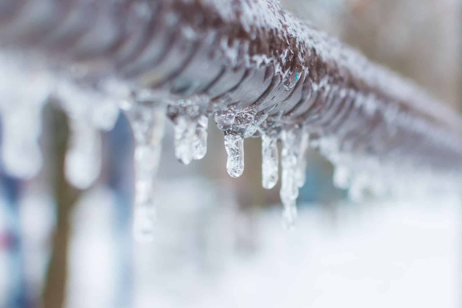 Close-up of a metal fence covered in icicles, with a snowy, blurred background. The scene captures the harsh winter weather—reminding homeowners to call Alpha Plumbing Heating and Cooling plumbers for reliable HVAC solutions during the cold.