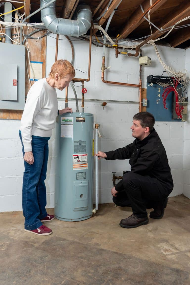 An Alpha Plumbing Heating and Cooling technician kneels next to a water heater, showing something to an older woman in a basement with exposed pipes, wires, and a cement floor.