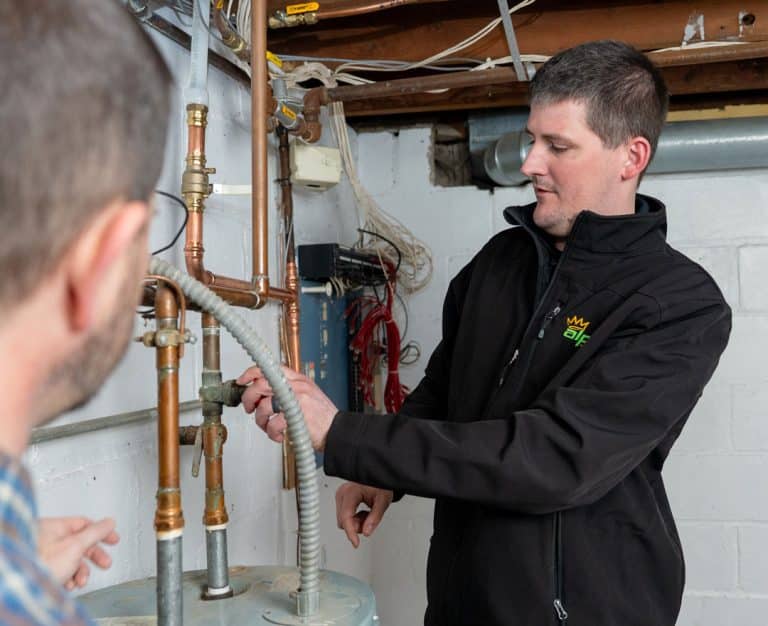 A technician in a black jacket adjusts pipes next to a water heater in a basement, while another person looks on. Exposed pipes and wires suggest Alpha Plumbing Heating and Cooling might also handle HVAC or furnace repair in this space.