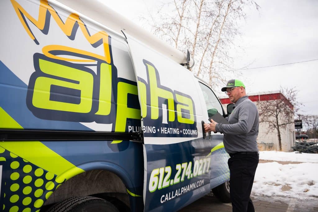 A worker in a green hat opens the side door of a colorful Alpha Plumbing, Heating, and Cooling van parked outside on a cloudy, snowy day. The van displays the company logo and contact info, highlighting their expert plumbers and HVAC services.