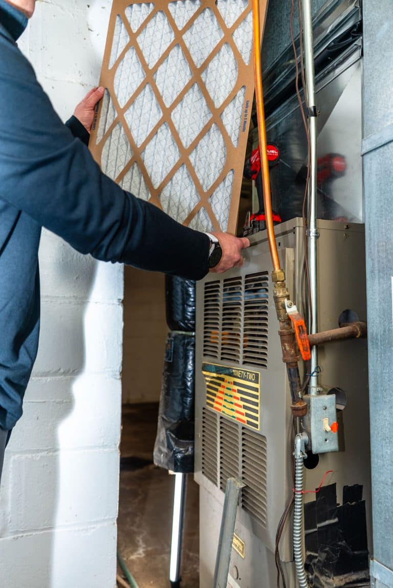 A person is installing or replacing a rectangular air filter in a furnace unit, part of an HVAC system, in a basement with white brick walls.