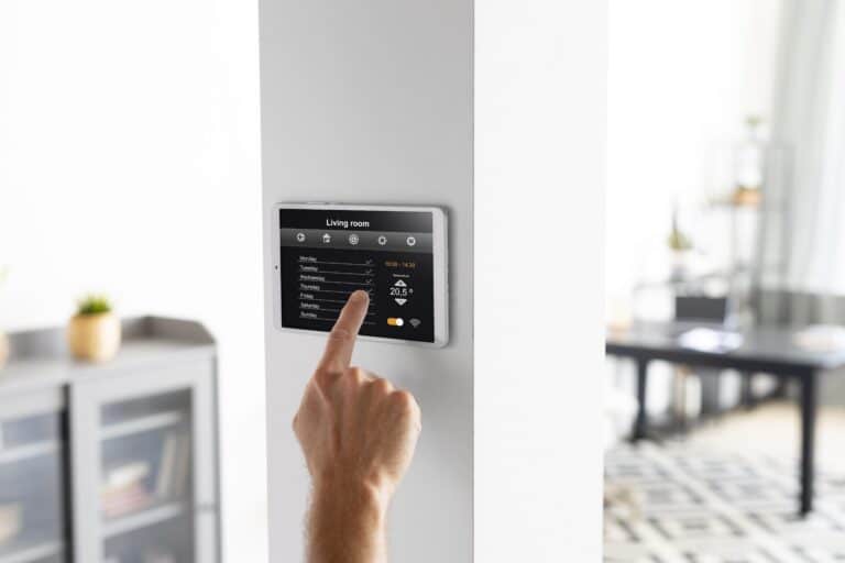 A person adjusts a smart home control panel mounted on a white wall, selecting the temperature for the living room—showcasing how smart thermostats can help answer the question: Are They Worth It for Minnesota Homes? A modern shelf and desk are visible in the background.