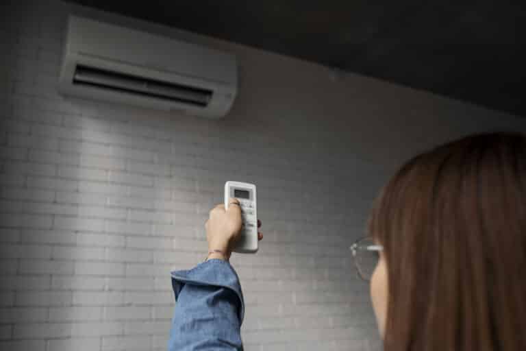 A person holding up a remote control and pointing it at a wall-mounted air conditioner on a white brick wall, emphasizing the importance of following a Spring HVAC Maintenance Checklist for Minnesota Homeowners.