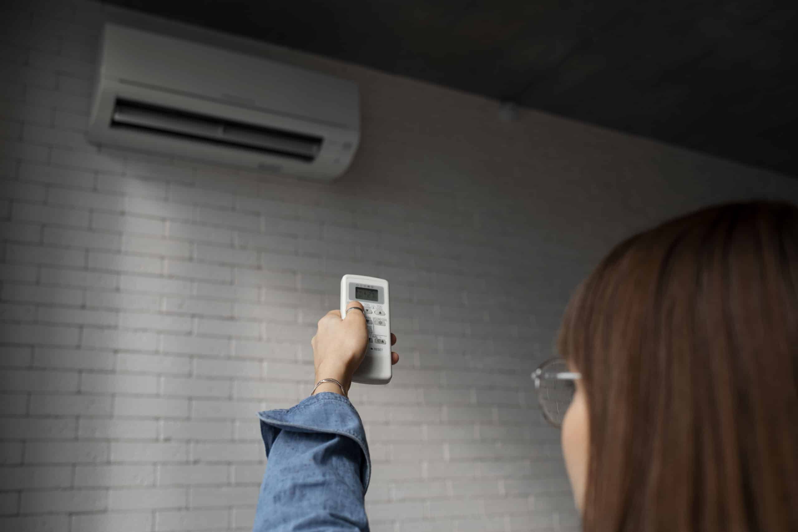 A person holding up a remote control and pointing it at a wall-mounted air conditioner on a white brick wall, emphasizing the importance of following a Spring HVAC Maintenance Checklist for Minnesota Homeowners.