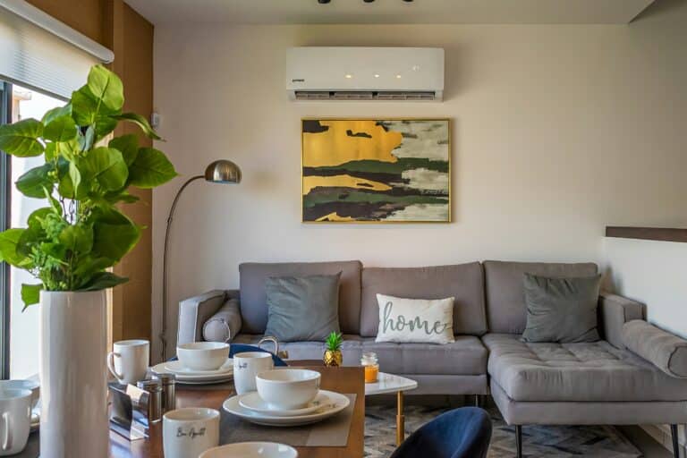 A modern living room with a gray sectional sofa, “home” pillow, abstract wall art, and a large potted plant. The air conditioner above the couch keeps things cool—a scene worthy of the Bloomington Air Conditioning Summer Guide.